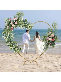 a man and woman holding hands in front of a heart shaped frame with flowers on the beach