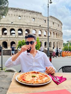 a man sitting at a table in front of an arena drinking from a bottle and eating pizza