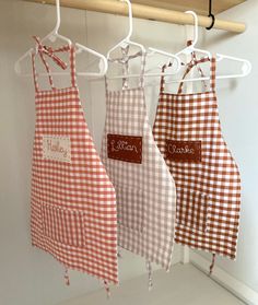 three aprons hanging on a rack with name tags attached to them, one is red and the other is white