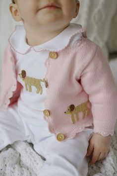 a baby wearing a pink sweater and white pants sitting on top of a knitted blanket