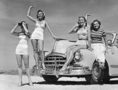 three women are posing in front of an old car with their arms up and one woman is holding her hat