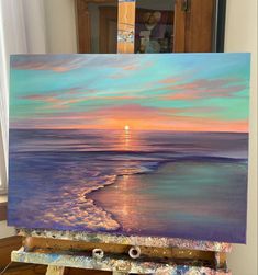 an easel with a painting on it that has a sunset in the background and water