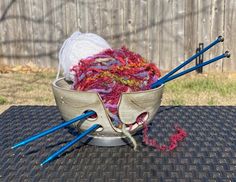 yarn and knitting needles sit in a bowl on a table outside, next to crochet hooks