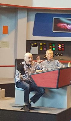 two men sitting on top of a chair in front of a tv screen with a red and blue boat