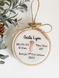 a cross stitch ornament hanging on a wall next to some plants and pine cones