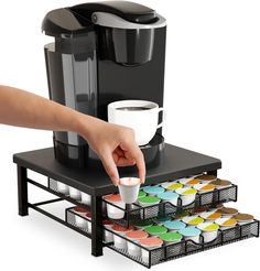 a coffee maker with cups on it and a person's hand reaching for the cup