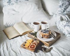 two cups of coffee, orange slices and waffles on a bed with an open book