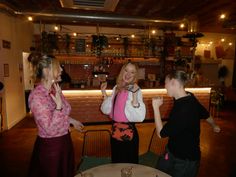 three women standing in front of a bar talking to each other while holding up their cell phones