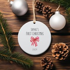 personalized first christmas ornament with red bow and pine cones on wooden background