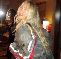 a woman with long blonde hair wearing a leather jacket and smiling at the camera while standing in front of a mirror