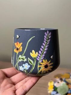 a hand holding up a painted cup with flowers on the outside and bees flying around it