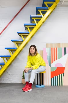 a woman is sitting on a bench in front of some stairs and paintings with bright colors