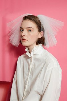 a woman with a veil on her head standing in front of a pink wall wearing a white shirt