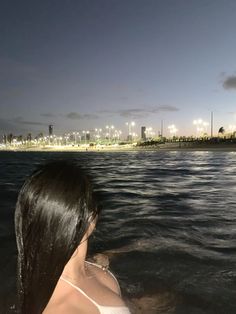 a woman sitting in the water looking out over the ocean at night with lights on