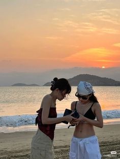 two young women standing on top of a sandy beach next to the ocean at sunset