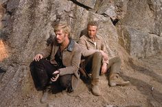 two men sitting on the ground next to large rocks with people in the background watching