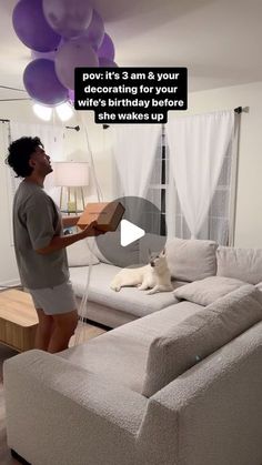 a woman holding a box in front of a couch with balloons on the ceiling and a dog laying on it