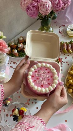 a person holding a heart shaped cake in front of a table full of flowers and desserts