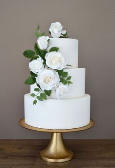 a three tiered cake with white flowers and green leaves on top is sitting on a gold plate