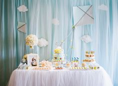 a table topped with lots of cakes and cupcakes next to a wall covered in clouds