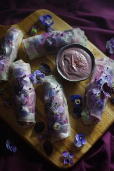 some food is sitting on a wooden board with purple flowers around it and a dipping sauce in a small bowl