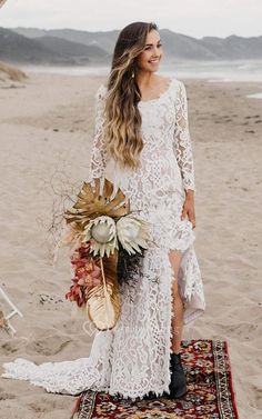 a woman standing on top of a rug next to a sandy beach holding a bouquet