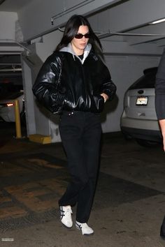 a woman in black leather jacket and white sneakers walking through an underground parking garage area