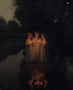 three women in white dresses holding candles standing in the water at night with trees around them