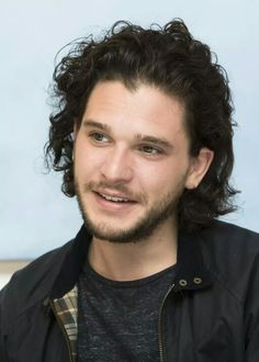 a man with long curly hair wearing a black jacket and looking at the camera while smiling