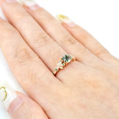 Inspired by nature's delicate beauty, the Moss Blossom ring features a round-cut natural moss agate as the centerpiece, symbolizing the serene connection between earth and art. The band is adorned with petal-like designs on each side, reminiscent of a blooming flower cradling a vibrant green fruit. Each petal is embellished with moissanite, adding a subtle sparkle that enhances the ring’s organic charm. The design seamlessly blends natural inspiration with elegance, perfect for those who appreciate understated sophistication in everyday wear. 𝗗𝗘𝗧𝗔𝗜𝗟𝗦𝗠𝗔𝗜𝗡 𝗦𝗧𝗢𝗡𝗘𝗚𝗲𝗺𝘀𝘁𝗼𝗻𝗲: Natural Moss Agate𝗖𝗼𝗹𝗼𝗿: Green𝗦𝗵𝗮𝗽𝗲: Round 5mm𝗖𝘂𝘁: Full𝗠𝗲𝗮𝘀𝘂𝗿𝗲𝗺𝗲𝗻𝘁: 1.0 ct approx𝗤𝘂𝗮𝗹𝗶𝘁𝘆: VS clarity grademoss agate are natural and can therefore slightly vary in color Nature-inspired Gemstone Flower Promise Ring, Nature-inspired Rose Gold Gemstone Rings, Nature-inspired Moss Agate Round Rings, Nature-inspired Moss Agate Rings, Delicate Flower Ring With Gemstone, Delicate Gemstone Flower Ring, Natural Inspiration, Trio Ring, Rutilated Quartz Ring