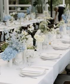 a long table is set with white and blue flowers in vases, plates and napkins