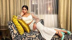 a woman sitting on top of a couch next to a window with curtains behind her