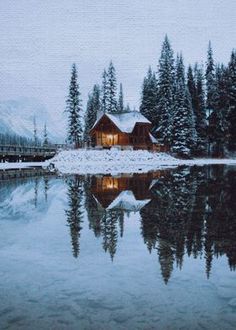 a cabin sits on the shore of a lake surrounded by snow covered mountains and pine trees