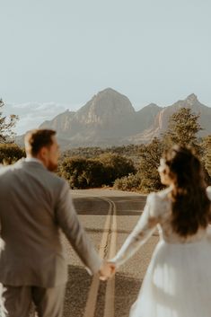 a bride and groom holding hands while walking down the road with mountains in the background