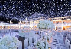 a banquet hall with tables and chairs covered in white flowers, lit up by fairy lights