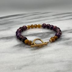 Introducing our meticulously handcrafted bracelet, a luxurious blend of deep, mesmerizing Purple Jasper and the captivating sheen of Gold Tiger Eye beads. This exquisite piece features 8mm Purple Jasper beads known for their grounding and protective properties, paired beautifully with 6mm Gold Tiger Eye beads that are believed to bring courage and confidence to the wearer. Each bead is delicately separated by elegant gold spacers, adding a touch of sophistication and timeless charm. The bracelet is securely fastened with a high-quality gold clasp, ensuring durability and ease of wear. Spiritual Amethyst Gold Bracelets, Gold Amethyst Beaded Bracelets For Jewelry Making, Gold Agate Beaded Bracelets With Gemstones, Gold Agate Gemstone Beaded Bracelets, Gold Agate Gemstone Beaded Bracelet, Gold Amethyst Bracelets For Healing, Gold Amethyst Beaded Bracelets, Gold Amethyst Jewelry With 8mm Beads, Gold Bracelets With Round Amethyst Beads