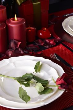 a white plate topped with a rose on top of a red table cloth next to candles