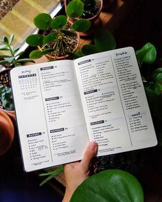 a person is holding an open book in their hand while sitting at a table with potted plants