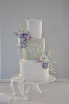 a three tiered cake with purple flowers on top and white ribbons around the edges