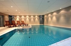 an indoor swimming pool with lounge chairs and lights on the wall above it, along with tiled flooring