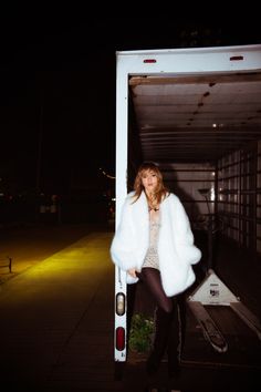 a woman standing in the back of a truck at night wearing a white fur coat