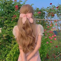 a woman with long blonde hair wearing a pink bow in front of some bushes and flowers