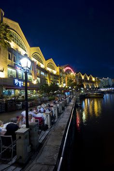 people are sitting at tables along the water in front of an illuminated shopping center and pier