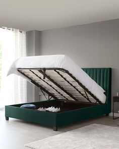 a green bed frame with white sheets and shoes underneath it in a living room next to a window