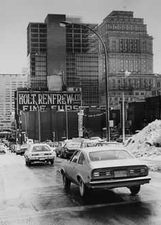 black and white photograph of cars driving down the street in front of tall buildings with signs on them