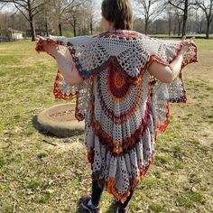 a woman wearing a crocheted shawl standing in a field with her arms outstretched