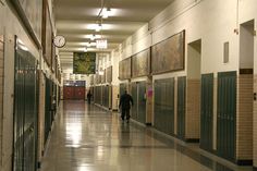 two people walking down an empty hallway in a building with several lockers on the walls