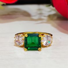 a green ring with three stones on it sitting on top of a white table next to a red rose