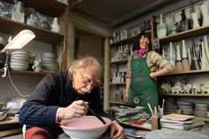 two women working in a pottery shop with bowls and plates on the shelves behind them