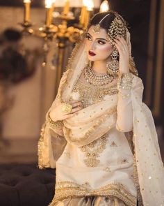 a woman in a white and gold bridal outfit sitting on a couch with candles behind her